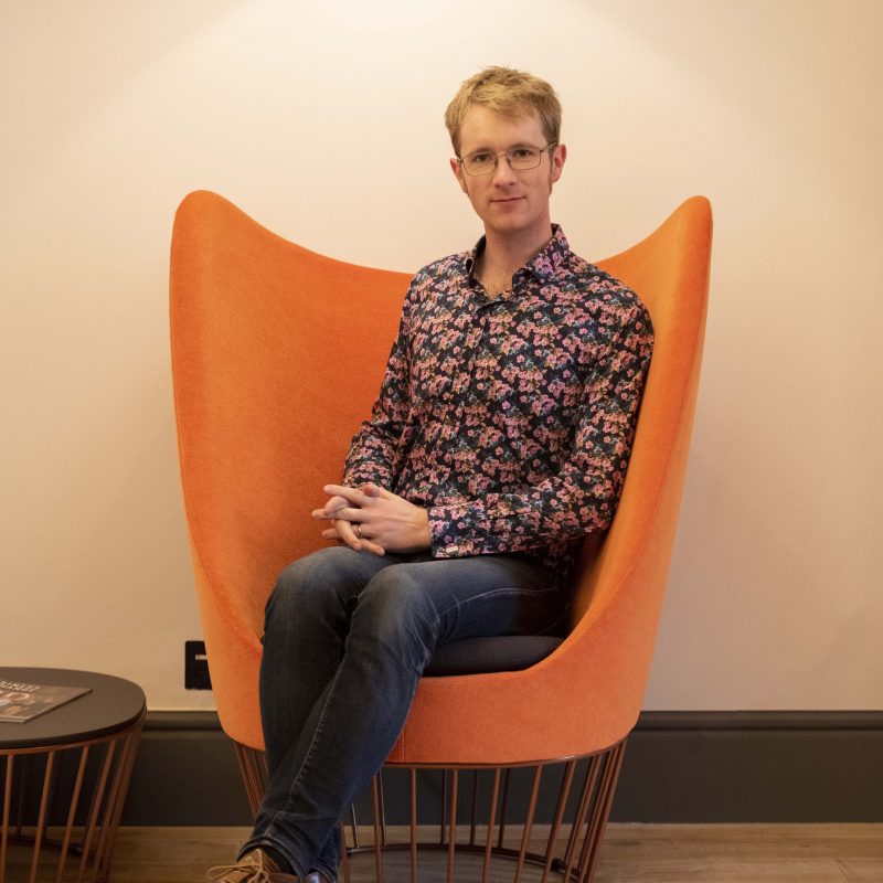 Moray Macdonald sitting in an orange chair, smiling at the camera. He is wearing glasses and a blue shirt with pink flowers.